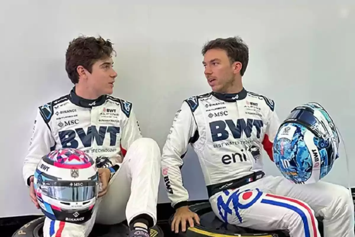 Two motorsport drivers in white BWT Alpine team suits are sitting across from each other, each holding their helmet and appearing to be conversing in a paddock setting. Two motorsport drivers in white BWT Alpine team suits are sitting across from each other, each holding their helmet and appearing to be conversing in a paddock setting.