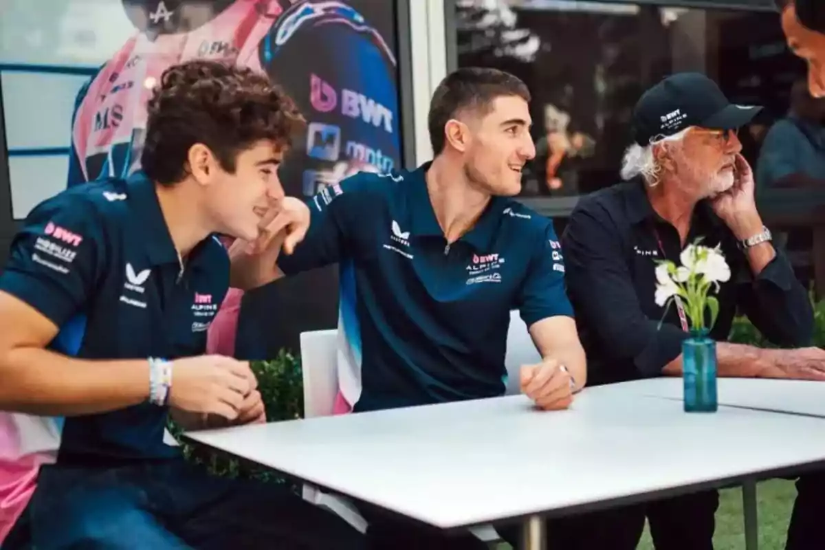 Three people sitting at a table, two of them wearing motorsport team jerseys and the third wearing a black cap and jersey, in an outdoor setting. Three people sitting at a table, two of them wearing motorsport team jerseys and the third wearing a black cap and jersey, in an outdoor setting.