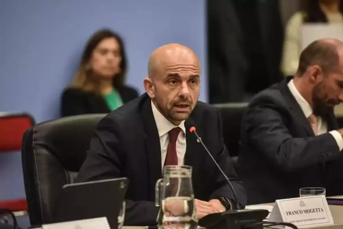 A bald man with a beard is speaking into a microphone during a formal meeting, dressed in a suit and tie, with people seated around him. A bald man with a beard is speaking into a microphone during a formal meeting, dressed in a suit and tie, with people seated around him.