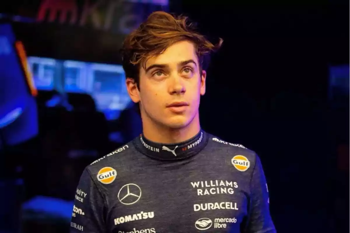 Un piloto de carreras con un uniforme azul oscuro de Williams Racing, con logotipos de patrocinadores como Mercedes, Duracell y Gulf, mirando hacia arriba en un entorno iluminado.