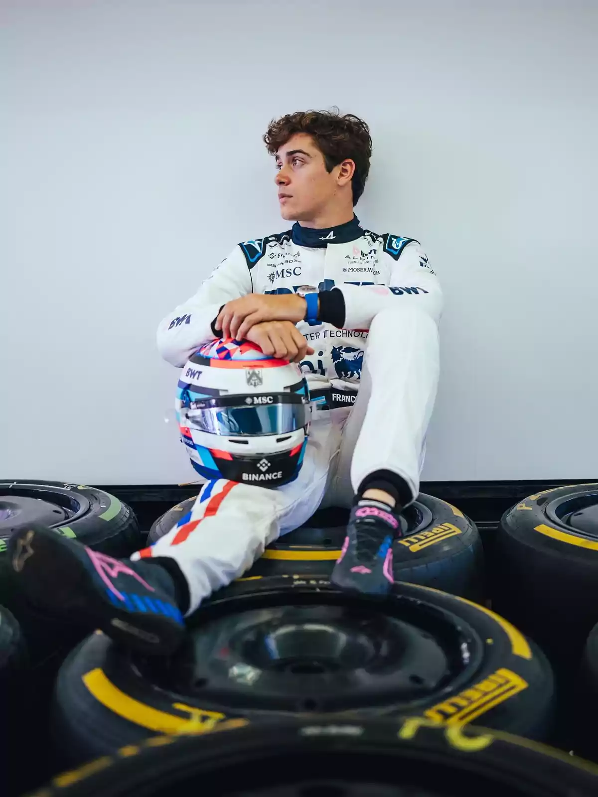 Piloto de automovilismo con traje blanco sentado sobre llantas de carreras sosteniendo un casco y mirando hacia un lado Piloto de automovilismo con traje blanco sentado sobre llantas de carreras sosteniendo un casco y mirando hacia un lado