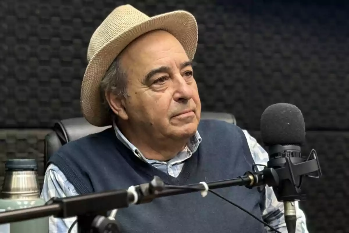 An older man with a hat is sitting in front of a microphone in a recording studio.
