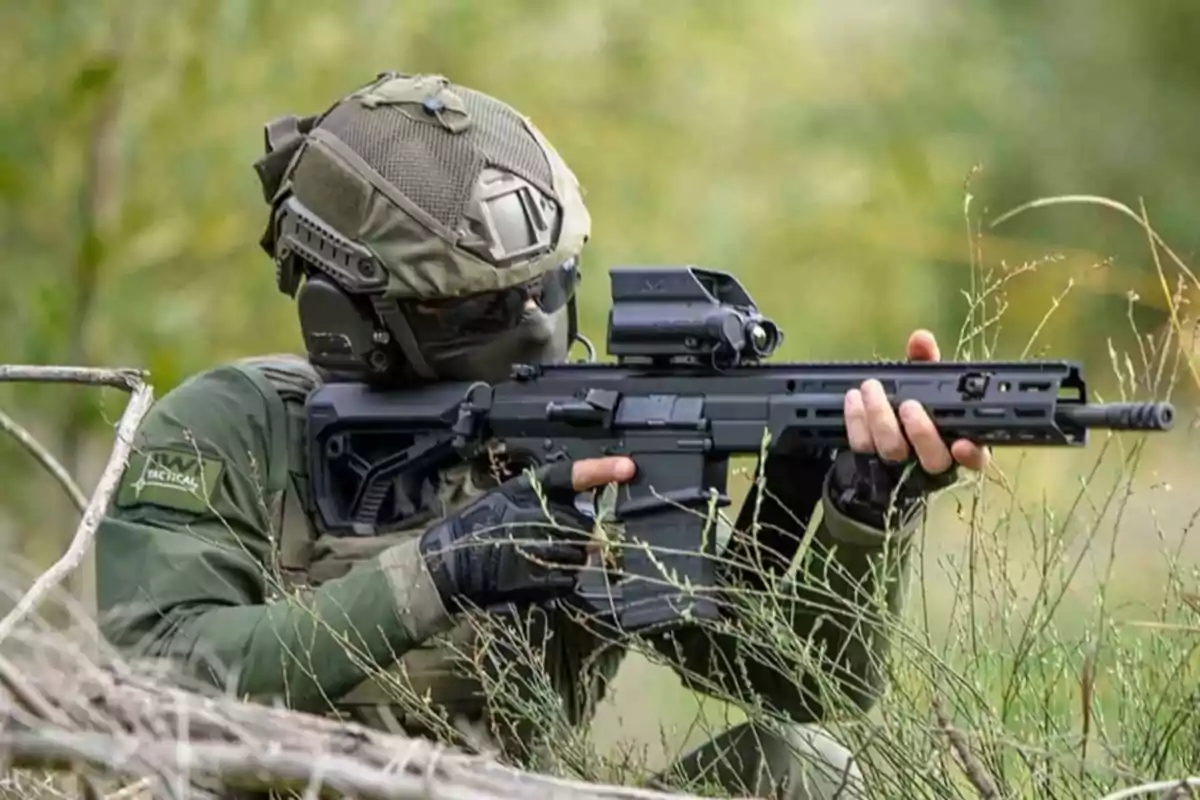 Un soldado con uniforme táctico y casco apunta un rifle en un entorno natural.