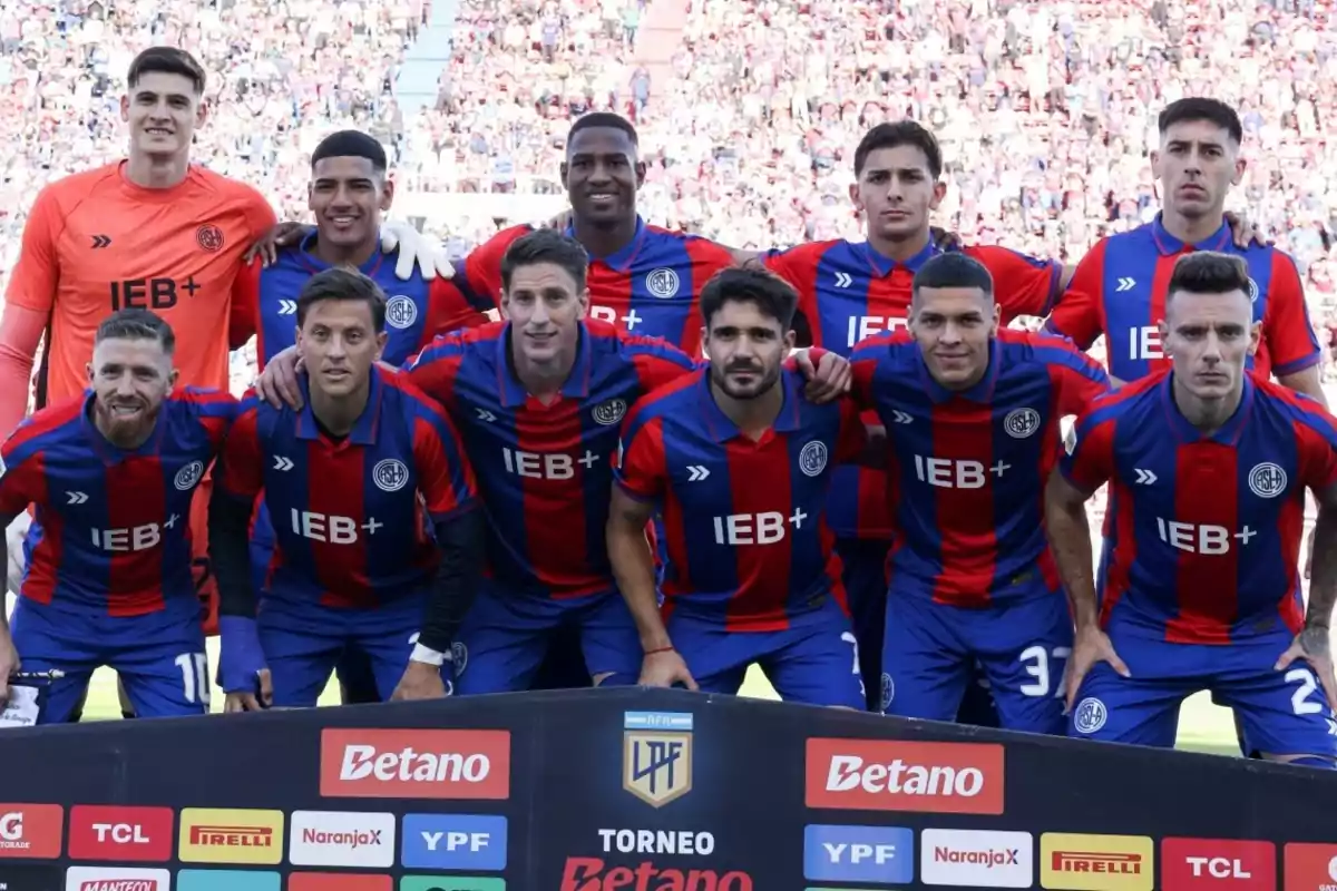 Un equipo de fútbol posando para una foto grupal en el campo de juego, con uniformes de rayas rojas y azules.