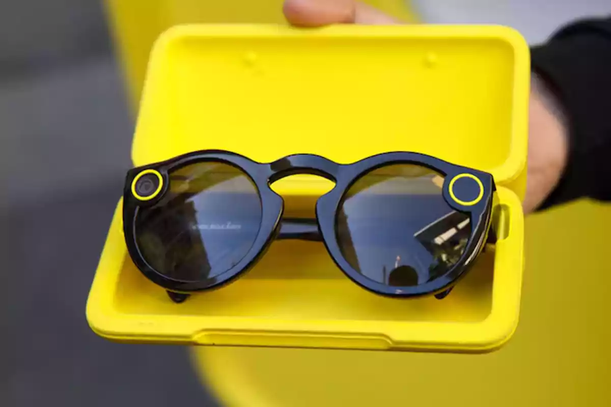 Gafas inteligentes negras con detalles amarillos dentro de un estuche amarillo sostenido por una mano Gafas inteligentes negras con detalles amarillos dentro de un estuche amarillo sostenido por una mano