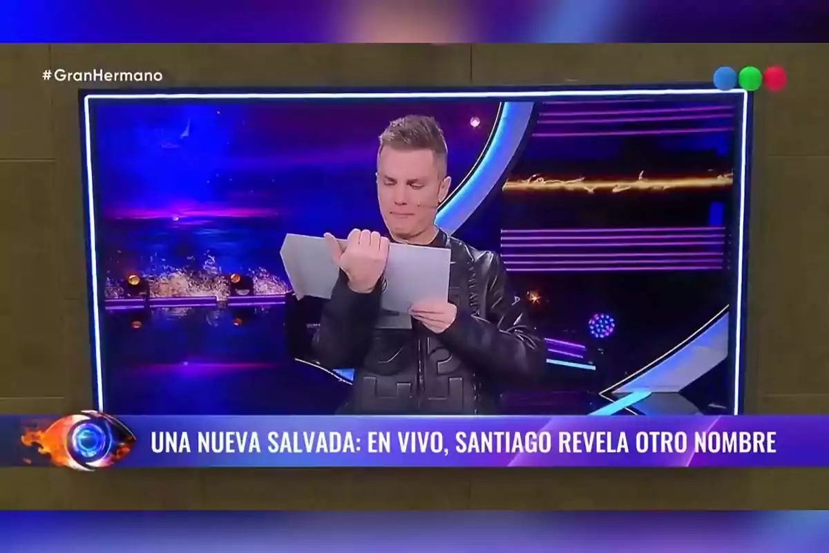Un presentador de televisión está en un set colorido, sosteniendo y leyendo un sobre mientras se transmite un programa en vivo. Un presentador de televisión está en un set colorido, sosteniendo y leyendo un sobre mientras se transmite un programa en vivo.