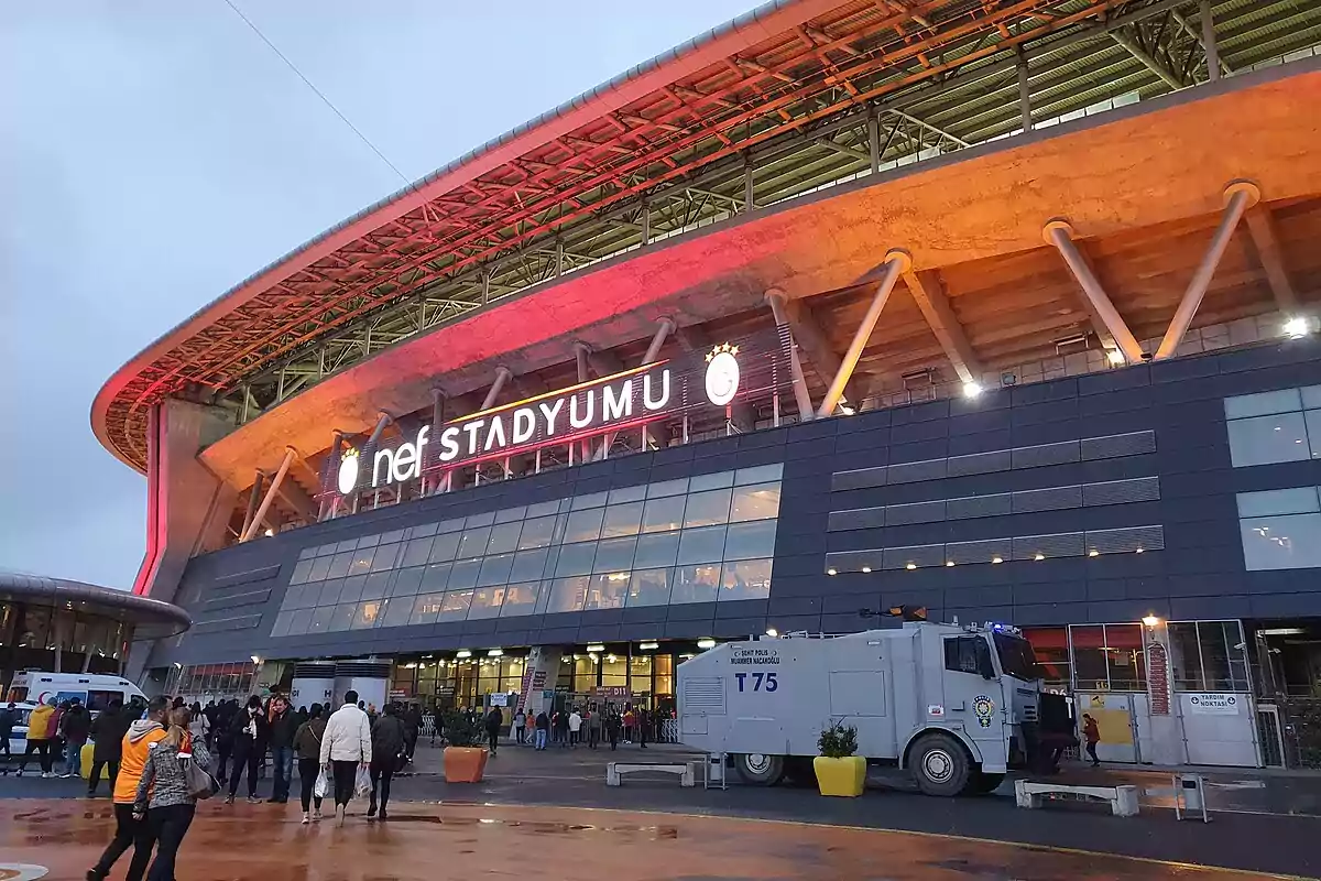 Personas caminando hacia la entrada de un estadio moderno con iluminación roja y un vehículo de seguridad estacionado al frente. Personas caminando hacia la entrada de un estadio moderno con iluminación roja y un vehículo de seguridad estacionado al frente.