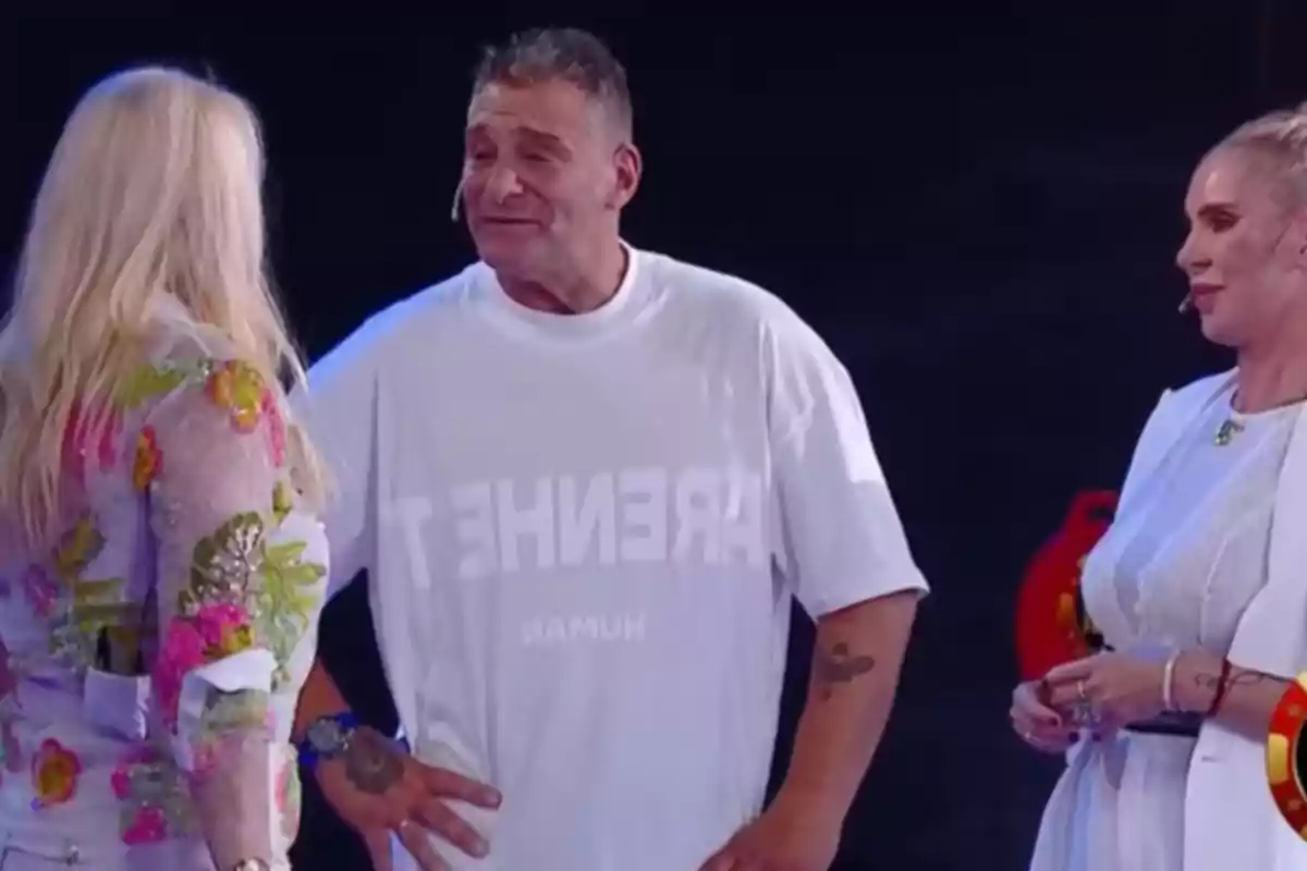 Tres personas conversando en un escenario, una de ellas lleva una camiseta blanca con texto. Tres personas conversando en un escenario, una de ellas lleva una camiseta blanca con texto.