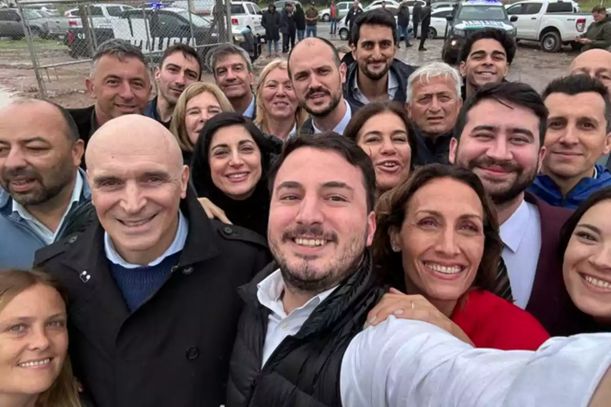 Un grupo de personas sonríe mientras se toma una selfie al aire libre, con vehículos y una cerca de fondo.