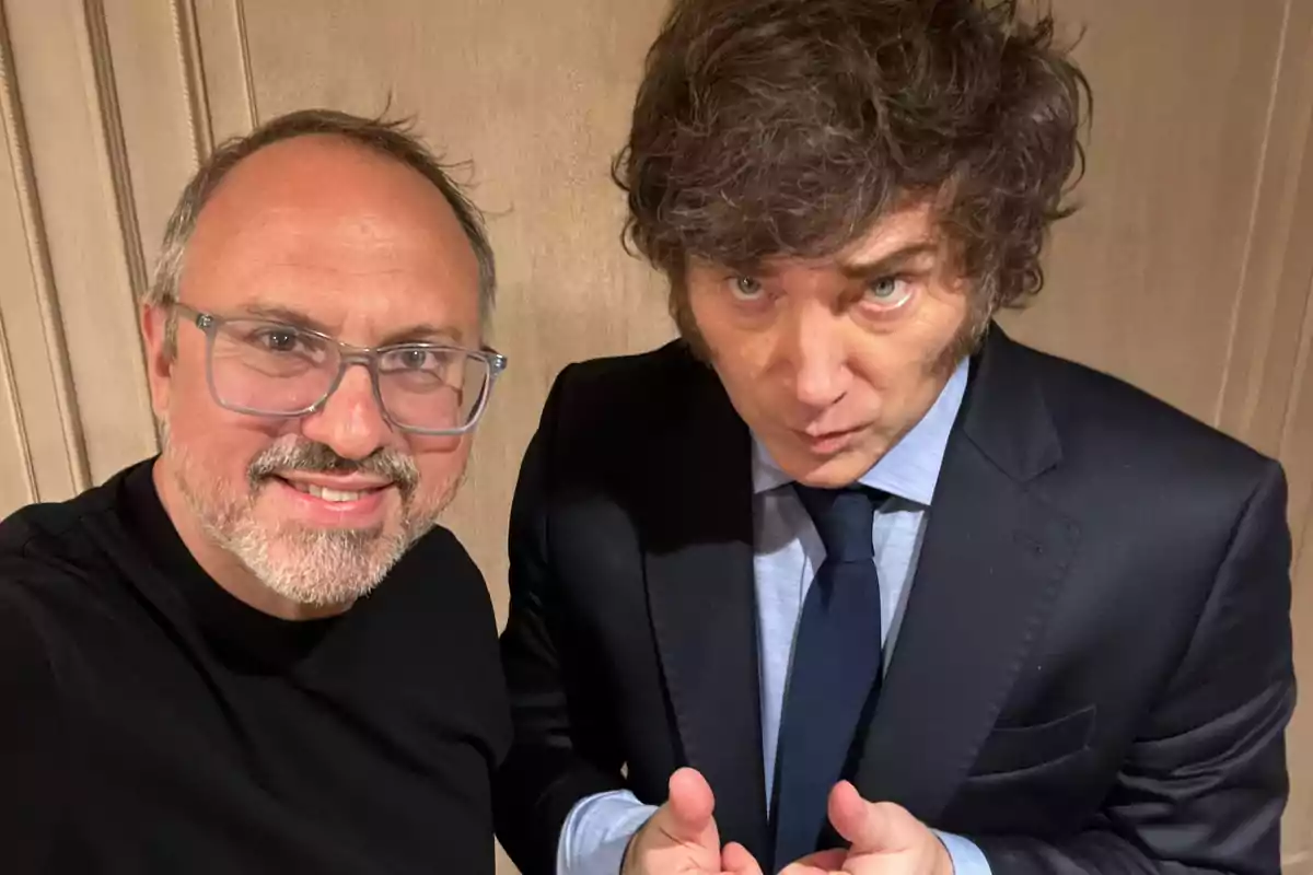 Dos hombres posan para una foto, uno lleva gafas y camiseta negra, el otro viste traje oscuro y corbata.