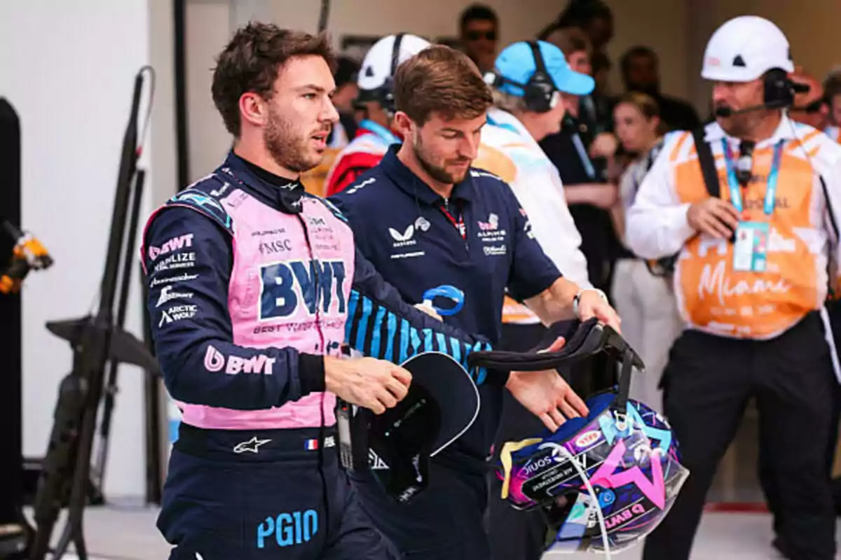A race car driver in a pink and blue suit walks alongside a team member at a motorsport event, while other people with headsets are in the background. A race car driver in a pink and blue suit walks alongside a team member at a motorsport event, while other people with headsets are in the background.
