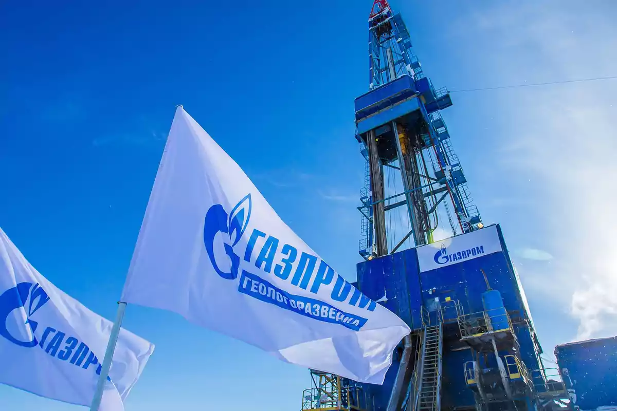 Banderas blancas con el logotipo de Gazprom ondean frente a una estructura de perforación azul bajo un cielo despejado.