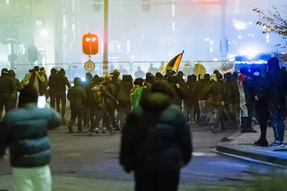 Un grupo de personas se reúne en una calle durante la noche con luces de vehículos y señales de tráfico visibles en el fondo.