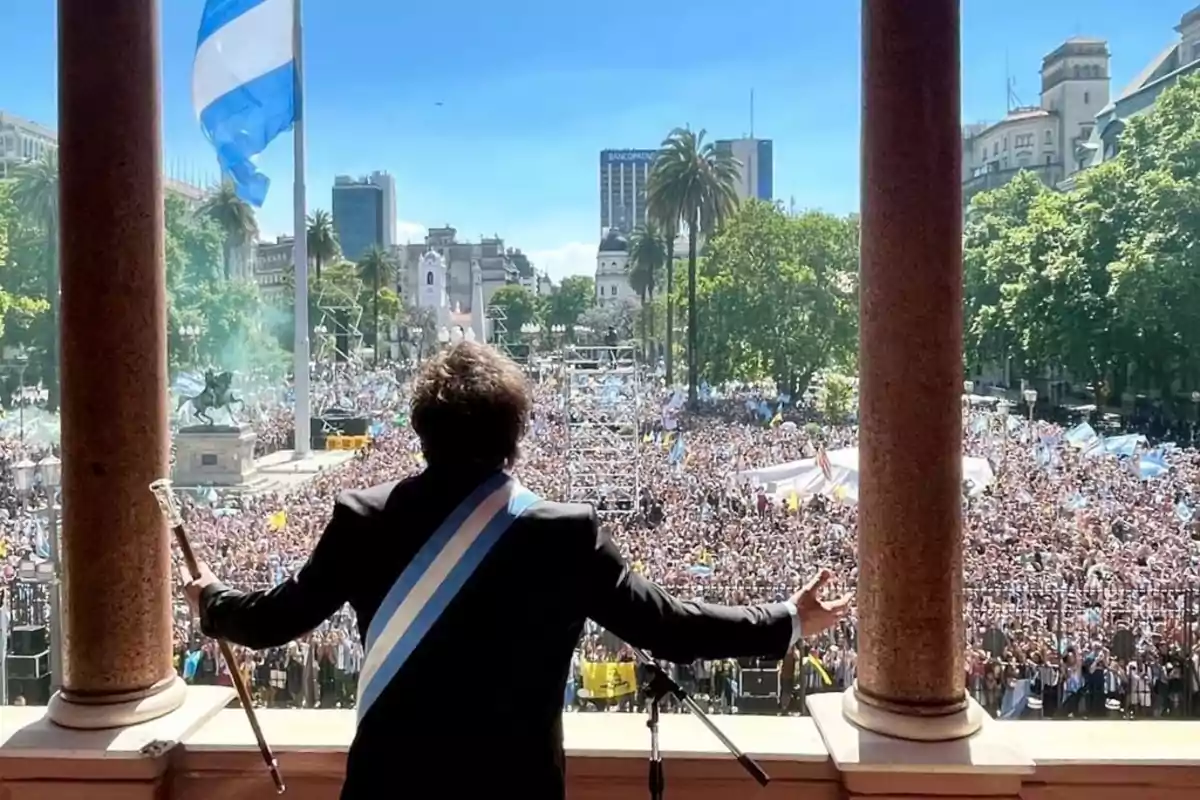 Una persona con una banda presidencial se dirige a una multitud desde un balcón, con una bandera ondeando y edificios al fondo.