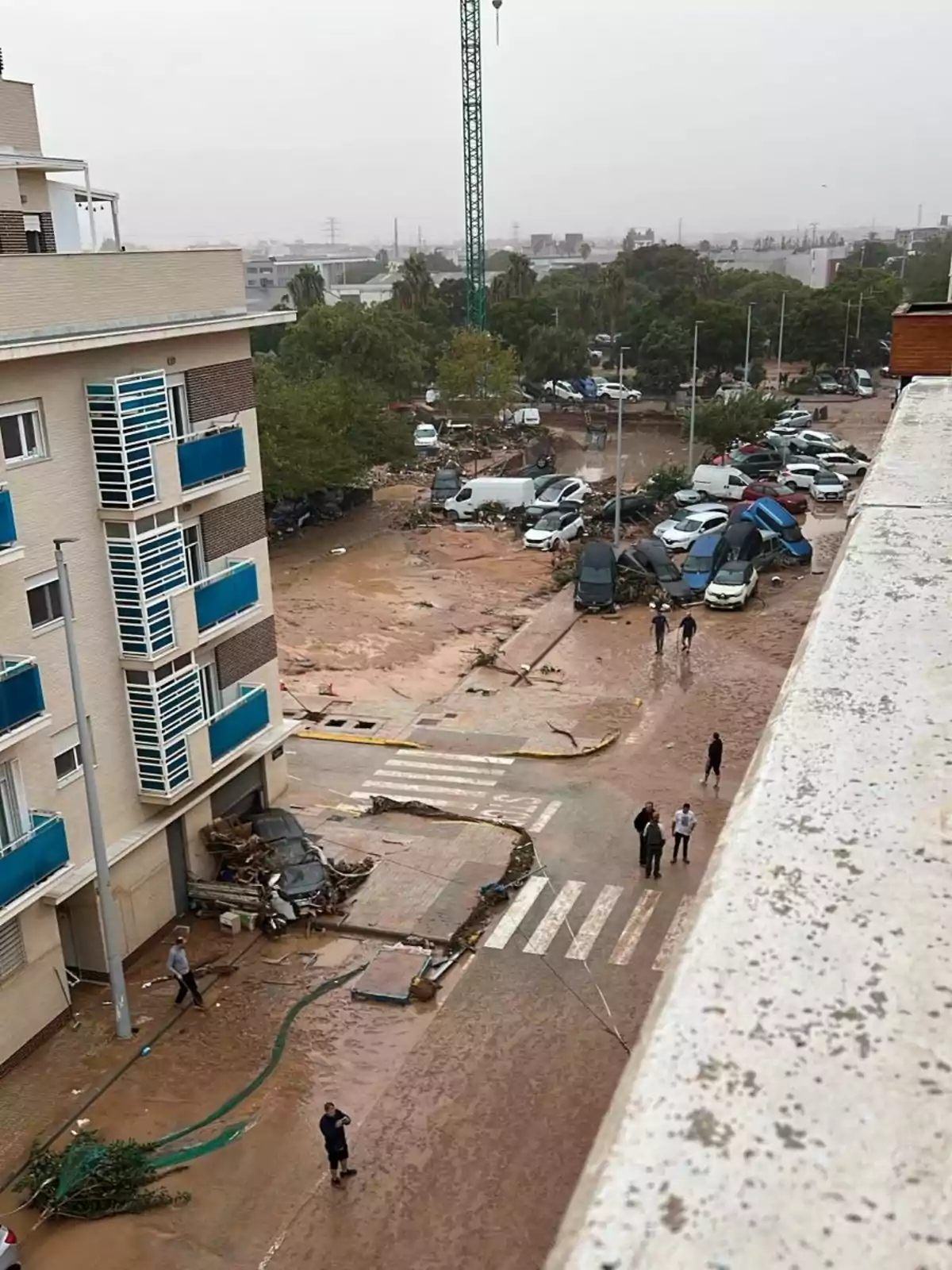 Vista aérea de una calle inundada con varios coches dañados y escombros, personas caminando por la zona afectada y un edificio residencial a la izquierda.