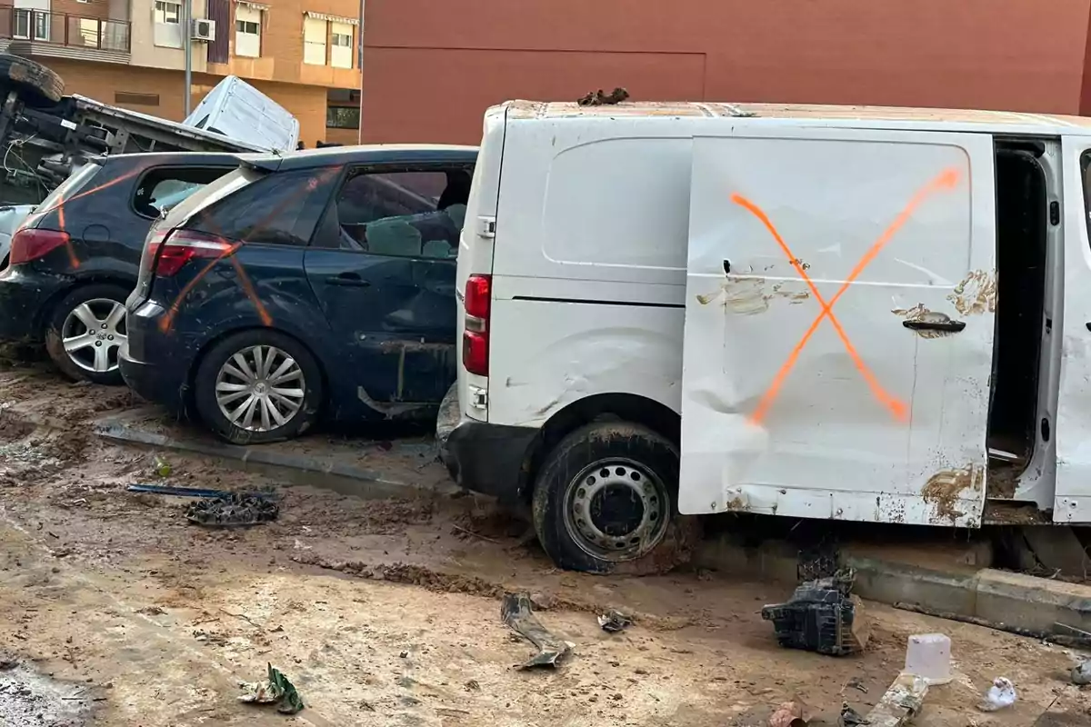 Vehículos dañados y cubiertos de barro con marcas de pintura en un área urbana después de un desastre.