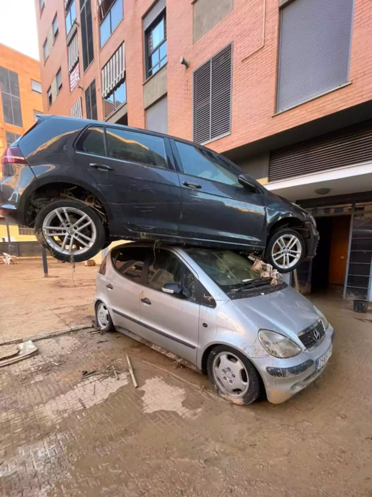 Dos coches apilados uno sobre otro en una calle con barro frente a un edificio de ladrillo.