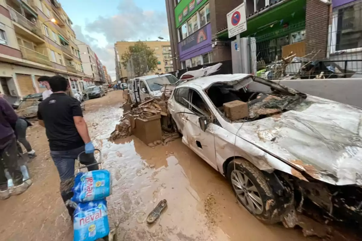 Calle inundada con coches dañados y personas caminando entre el barro llevando botellas de agua.