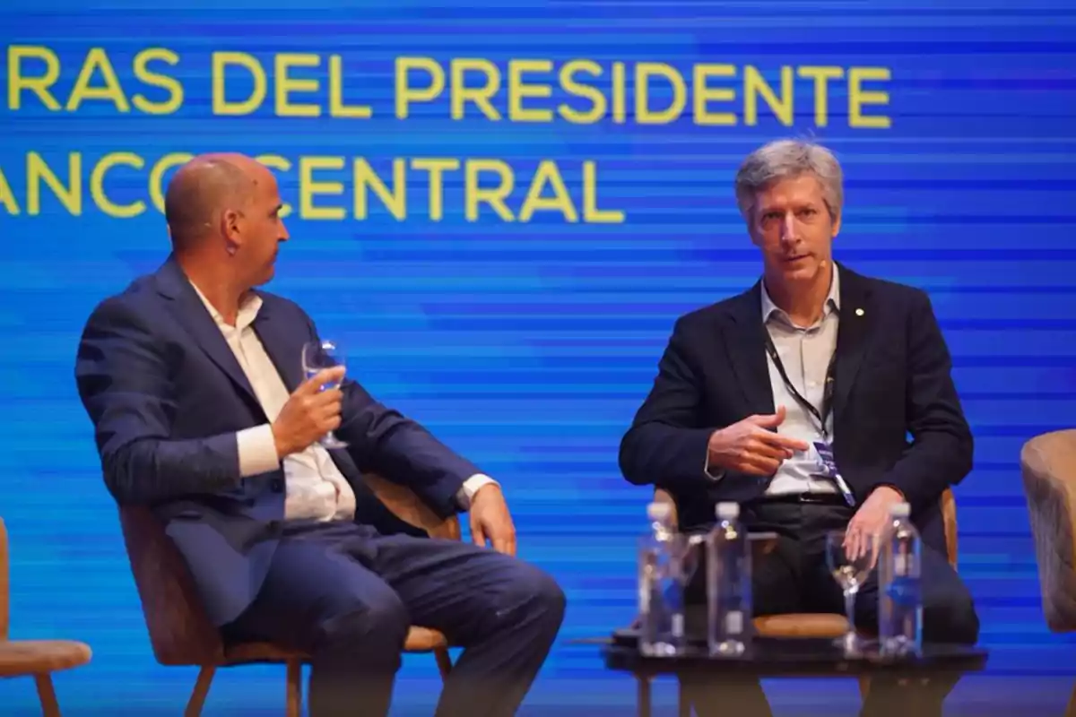 Dos hombres en un escenario con fondo azul y texto amarillo, uno de ellos sostiene una copa.