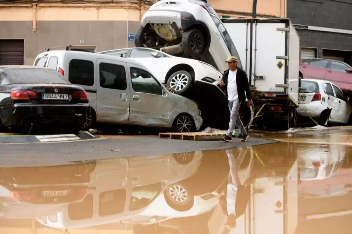 Un hombre camina junto a varios coches apilados y dañados tras una inundación, con charcos de agua en el suelo.