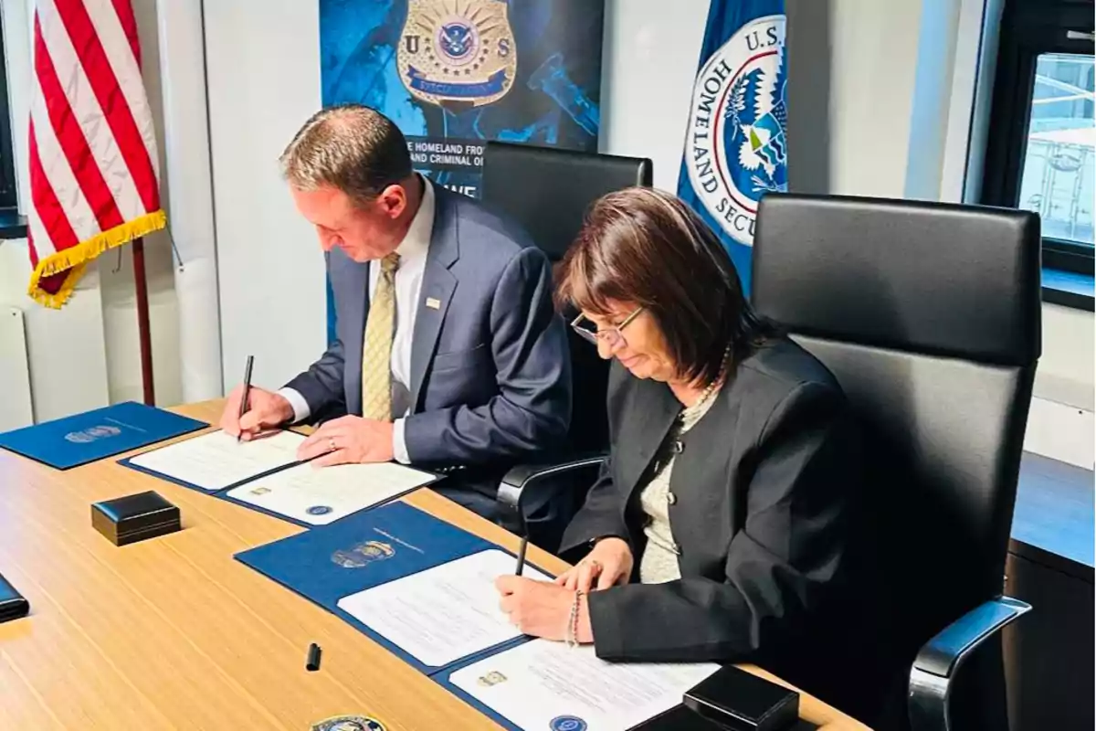 Dos personas sentadas en una mesa firmando documentos oficiales con banderas y emblemas gubernamentales en el fondo.