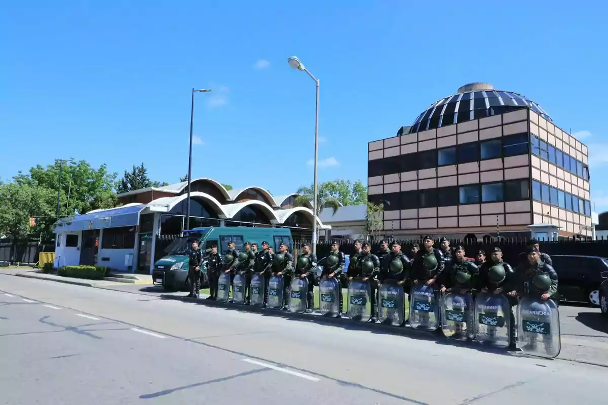 Un grupo de policías con equipo antidisturbios se encuentra alineado frente a un edificio moderno y un vehículo policial en una calle despejada bajo un cielo azul.