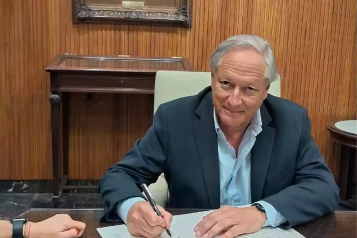 Un hombre de cabello canoso y traje oscuro está sentado en una mesa firmando un documento.