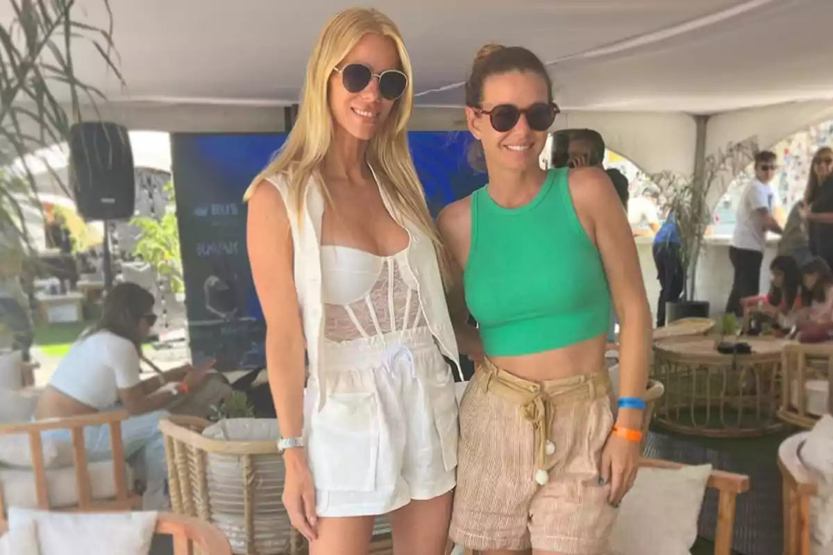 Two women smile and pose together in an outdoor setting with wicker decorations and plants; both are wearing sunglasses and summer clothes, one in a white outfit and the other in a green top and beige shorts. Two women smile and pose together in an outdoor setting with wicker decorations and plants; both are wearing sunglasses and summer clothes, one in a white outfit and the other in a green top and beige shorts.