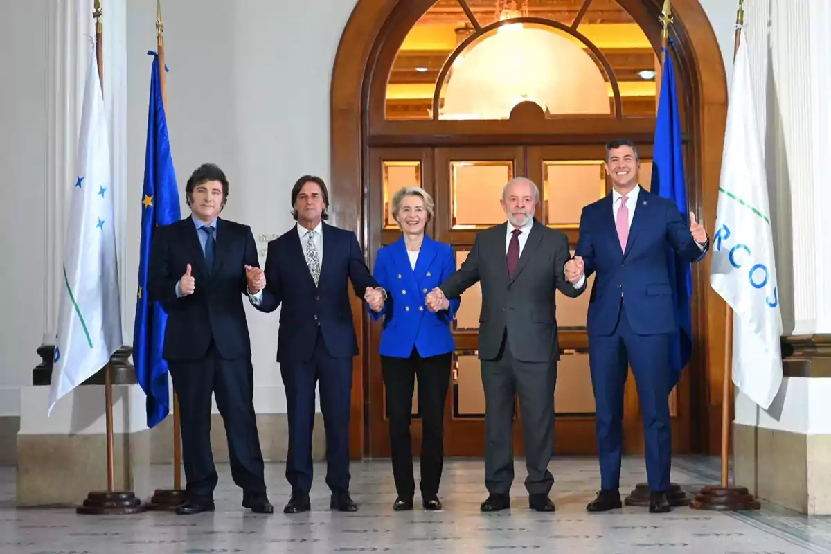 Cinco personas de pie, tomadas de la mano, frente a una puerta de madera, con banderas a los lados.