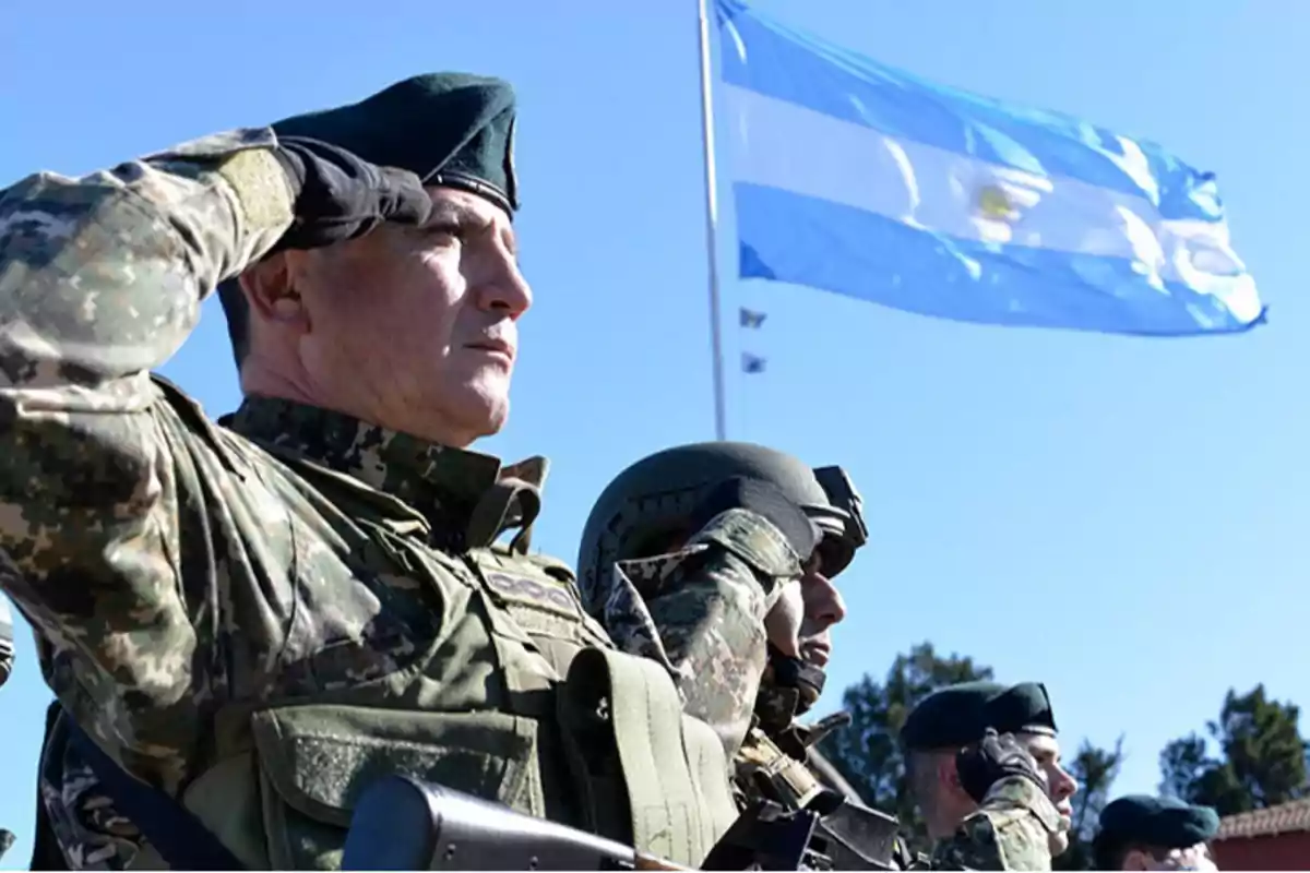 Soldados en uniforme militar saludando frente a una bandera de Argentina ondeando en el fondo.