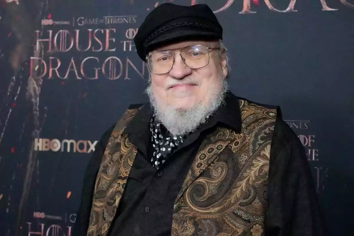 White-bearded man with glasses and a black cap posing in front of a House of the Dragon promotional backdrop White-bearded man with glasses and a black cap posing in front of a House of the Dragon promotional backdrop