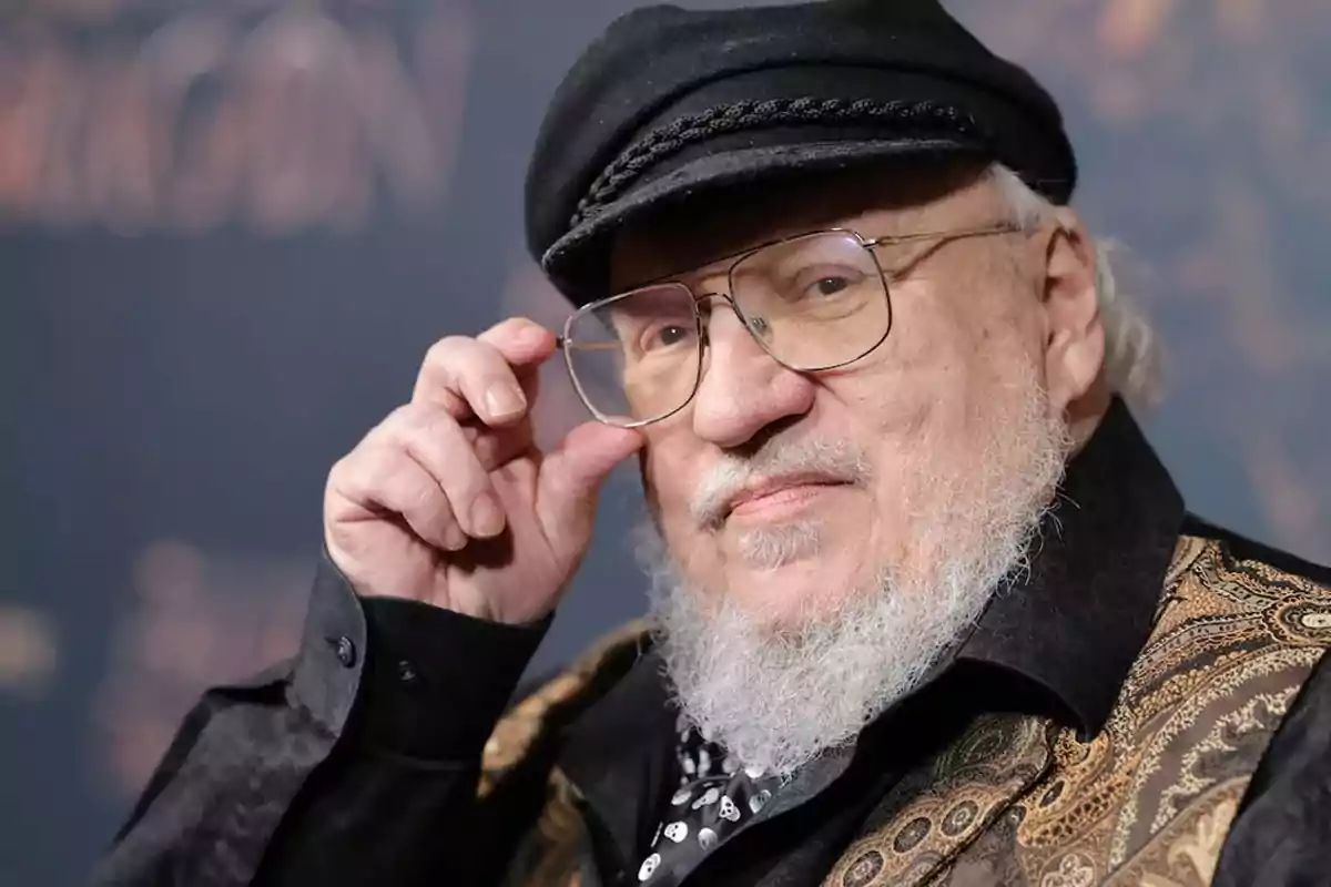 Un hombre con barba blanca y gafas, vistiendo una gorra negra y una chaqueta con diseño detallado, mira a la cámara mientras ajusta sus gafas. Un hombre con barba blanca y gafas, vistiendo una gorra negra y una chaqueta con diseño detallado, mira a la cámara mientras ajusta sus gafas.