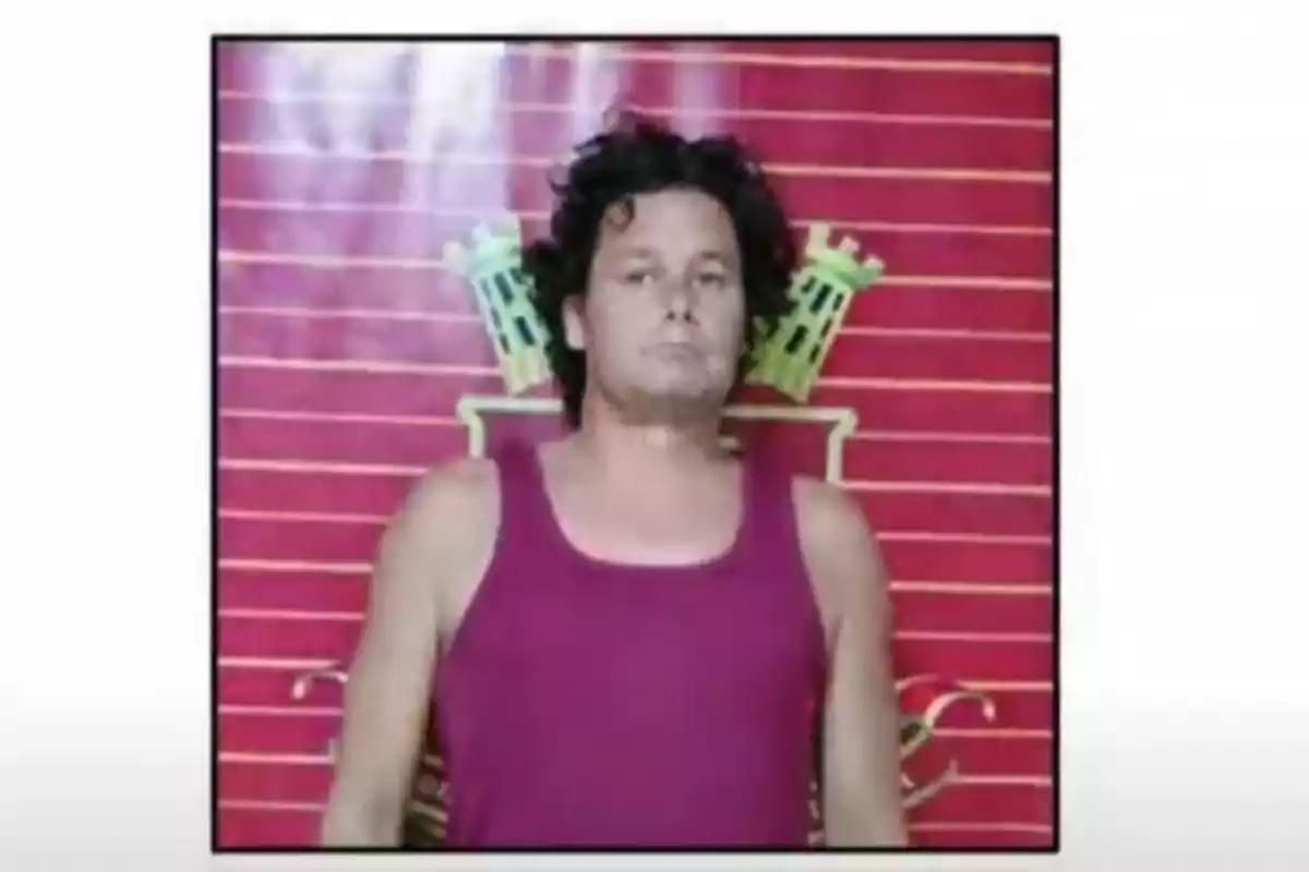 A man in a dark red tank top is standing in front of a red background with horizontal lines and a design in the center.
