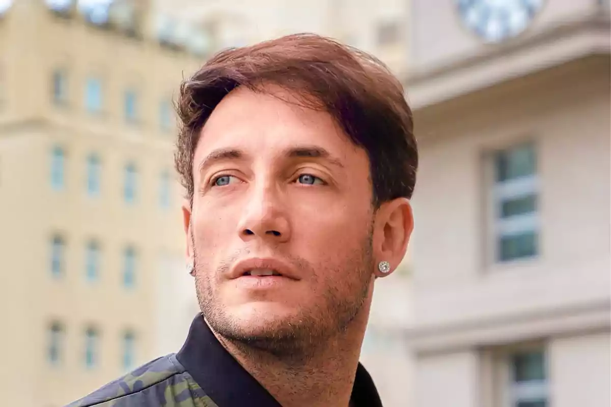 Hombre joven con barba corta y aretes mirando hacia un lado con edificios de fondo