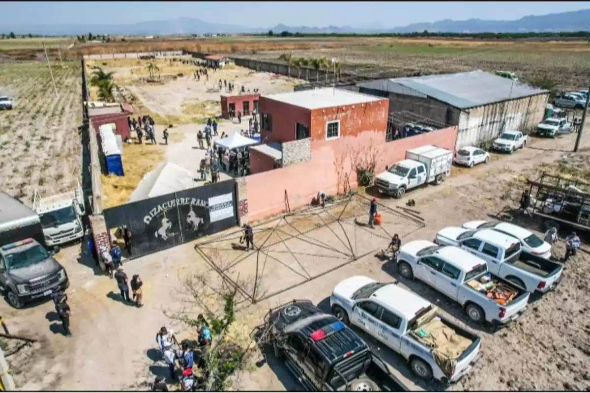 Vista aérea de un rancho con varios vehículos estacionados y personas reunidas en el área central, rodeado de campos y montañas al fondo. Vista aérea de un rancho con varios vehículos estacionados y personas reunidas en el área central, rodeado de campos y montañas al fondo.