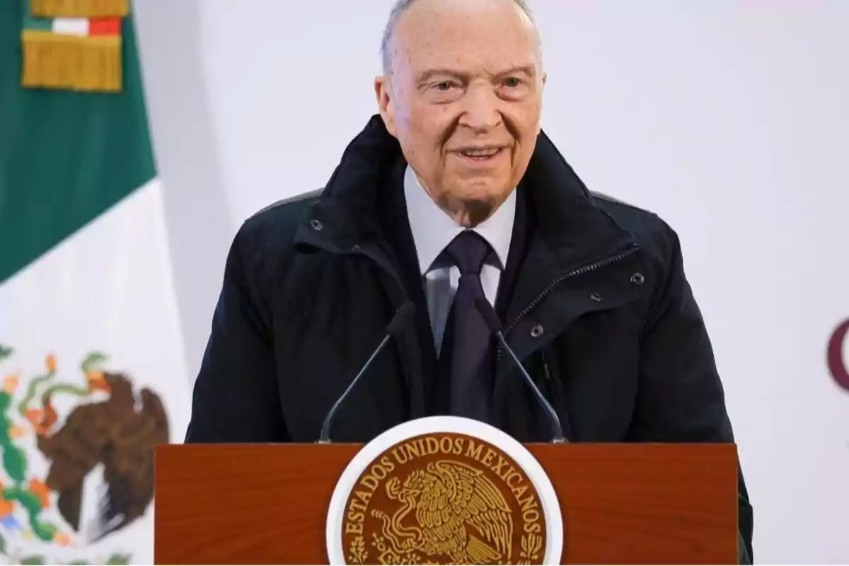Un hombre de pie detrás de un podio con el escudo de México y una bandera mexicana al fondo. Un hombre de pie detrás de un podio con el escudo de México y una bandera mexicana al fondo.