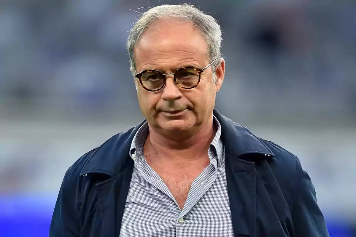 A man with glasses and gray hair is wearing a dark jacket and a checkered jersey, with a blurred background. A man with glasses and gray hair is wearing a dark jacket and a checkered jersey, with a blurred background.