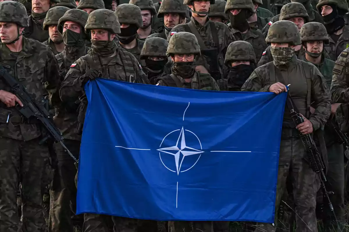 Soldiers in camouflage uniforms hold a blue NATO flag. Soldiers in camouflage uniforms hold a blue NATO flag.