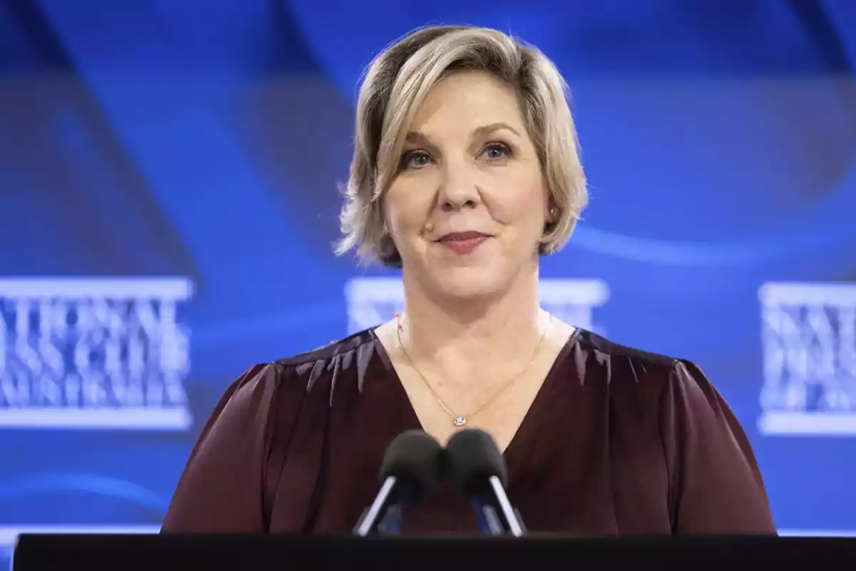 Blonde short-haired woman speaking in front of a microphone with a blue background. Blonde short-haired woman speaking in front of a microphone with a blue background.