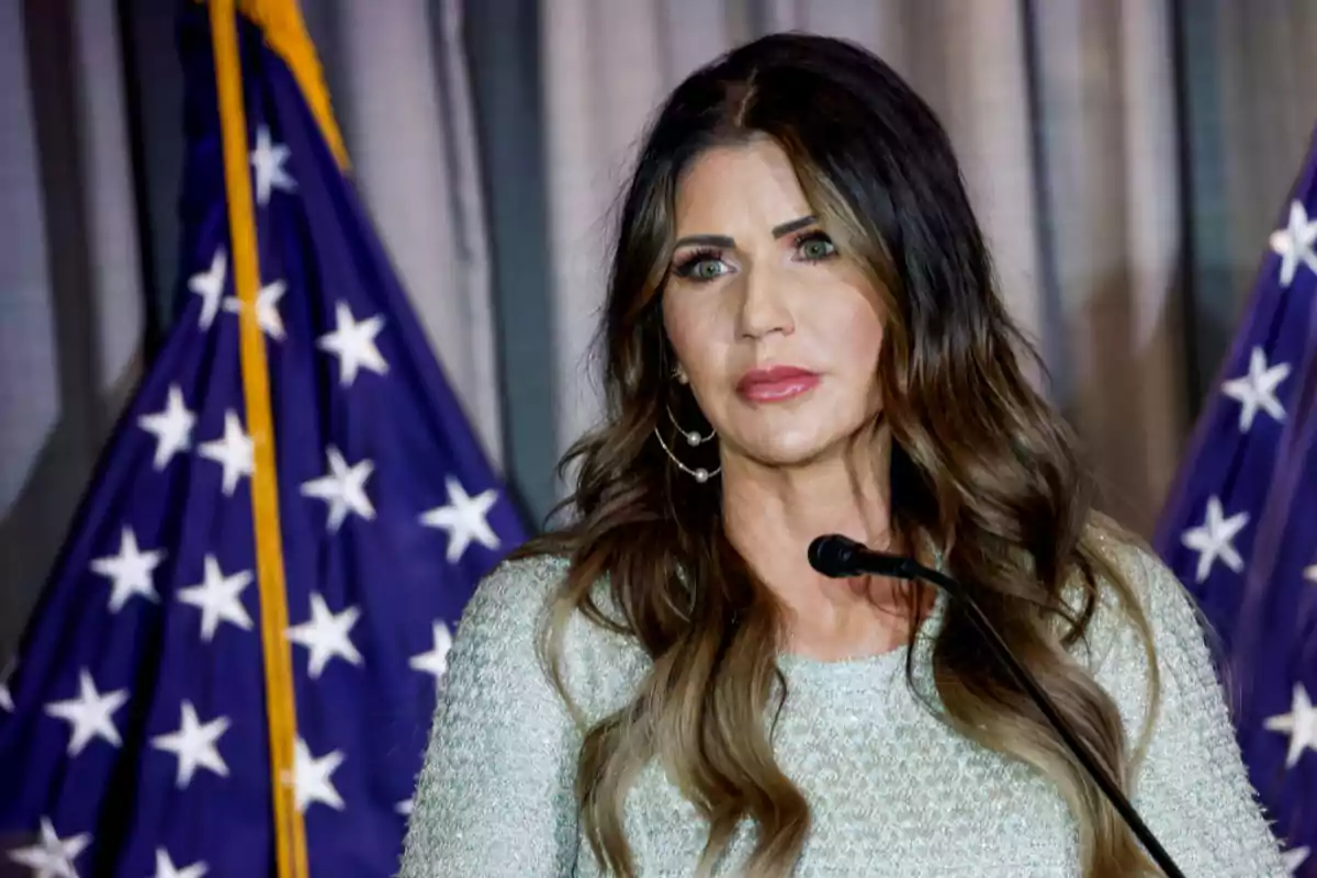 A woman with long dark hair speaks in front of a microphone with flags in the background. A woman with long dark hair speaks in front of a microphone with flags in the background.