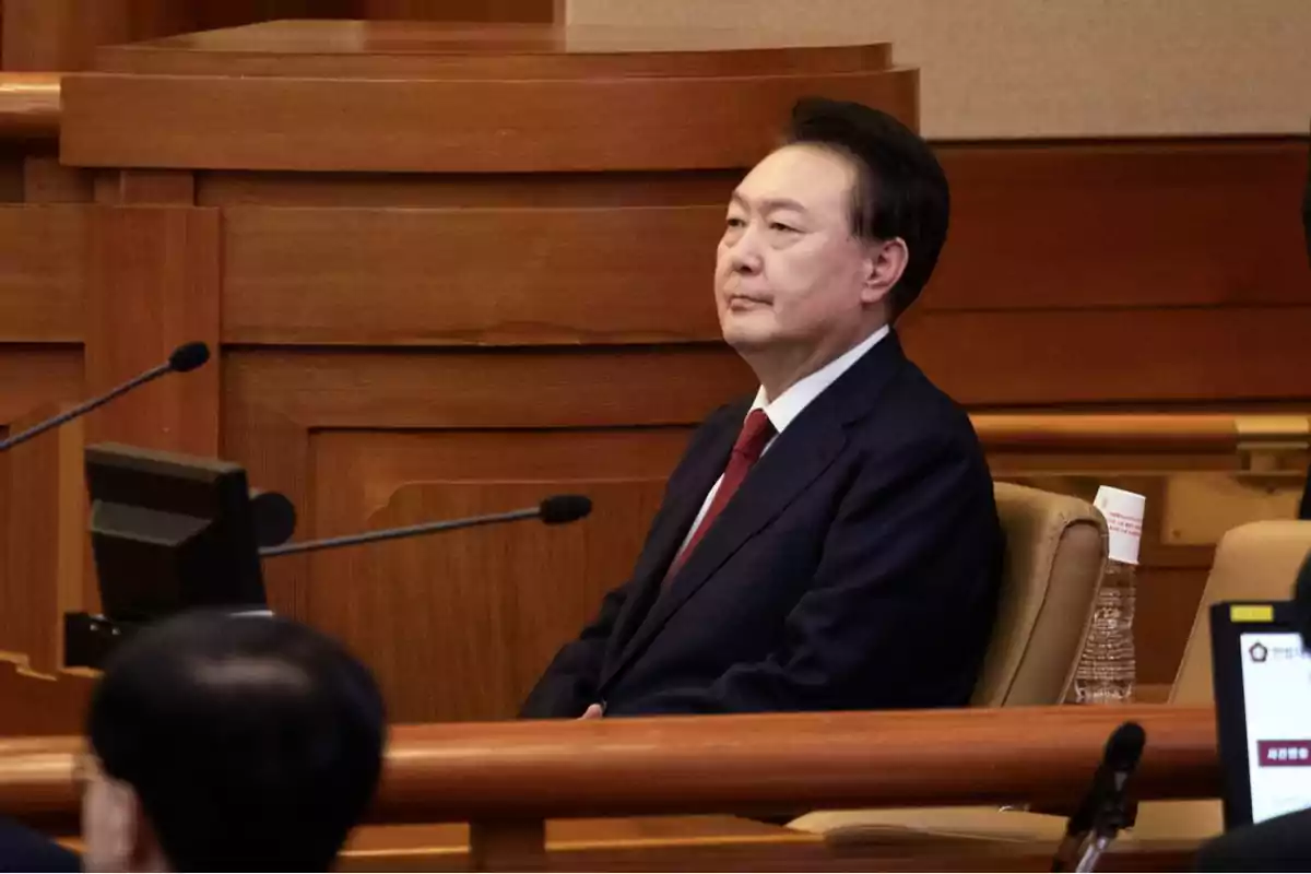 A man in a dark suit and red tie is sitting in a wooden conference room, in front of a microphone and a screen. A man in a dark suit and red tie is sitting in a wooden conference room, in front of a microphone and a screen.