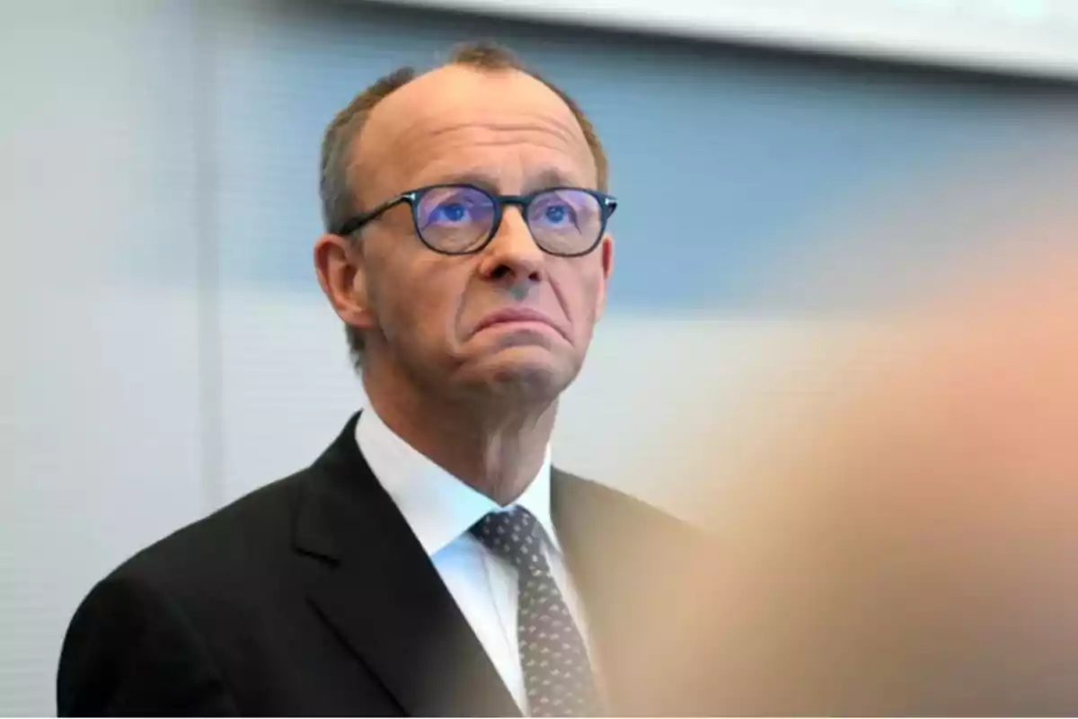 Un hombre con gafas y traje oscuro mira hacia arriba con expresión seria en un entorno profesional. Un hombre con gafas y traje oscuro mira hacia arriba con expresión seria en un entorno profesional.