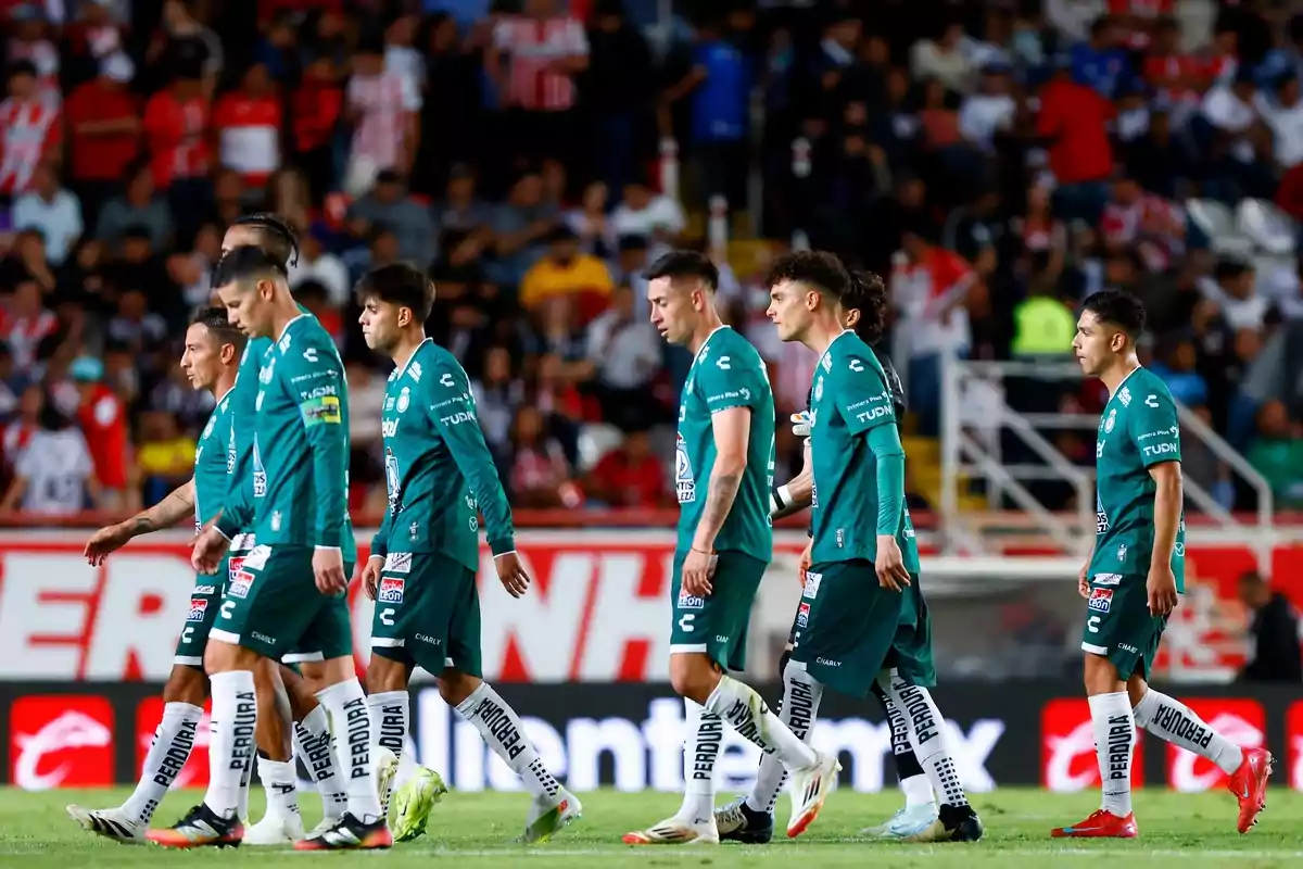Soccer players in green uniforms walking on the field during a match, with spectators in the stands in the background.