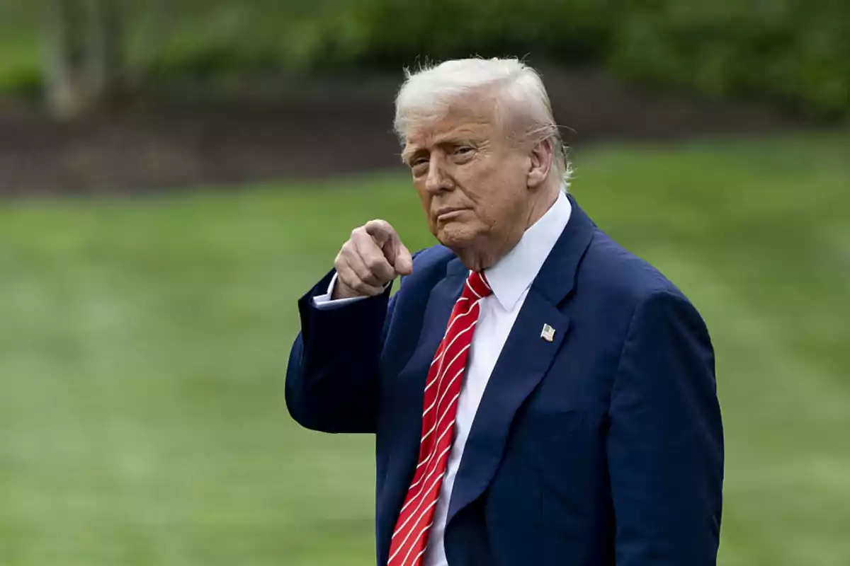 Blond-haired man in a blue suit and red tie pointing forward in an outdoor setting with grass in the background