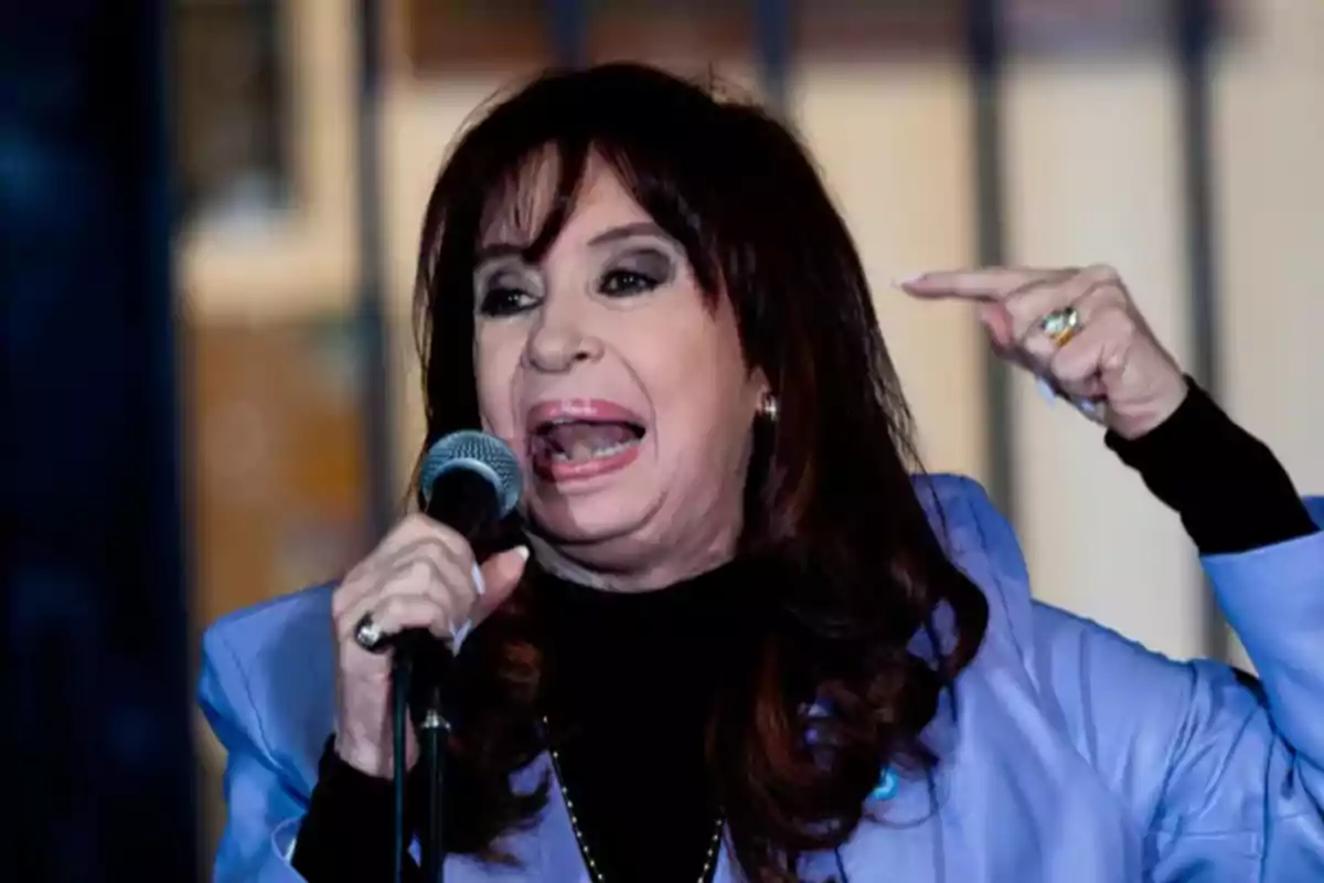 Mujer de cabello oscuro hablando en un micrófono y gesticulando con la mano mientras usa un saco celeste