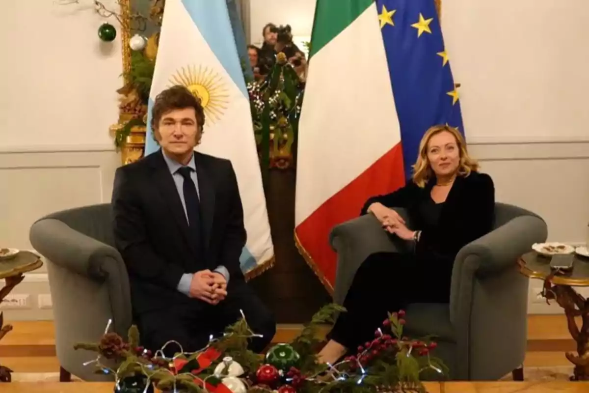 Dos personas sentadas en sillones frente a banderas de Argentina, Italia y la Unión Europea, con decoraciones navideñas alrededor.