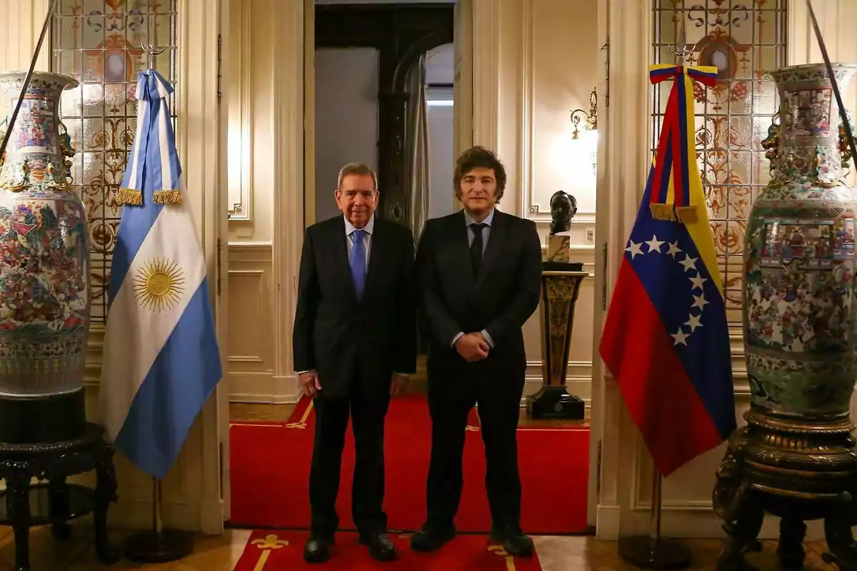 Dos hombres de traje posan en un elegante salón con alfombra roja y banderas de Argentina y Venezuela a los lados.