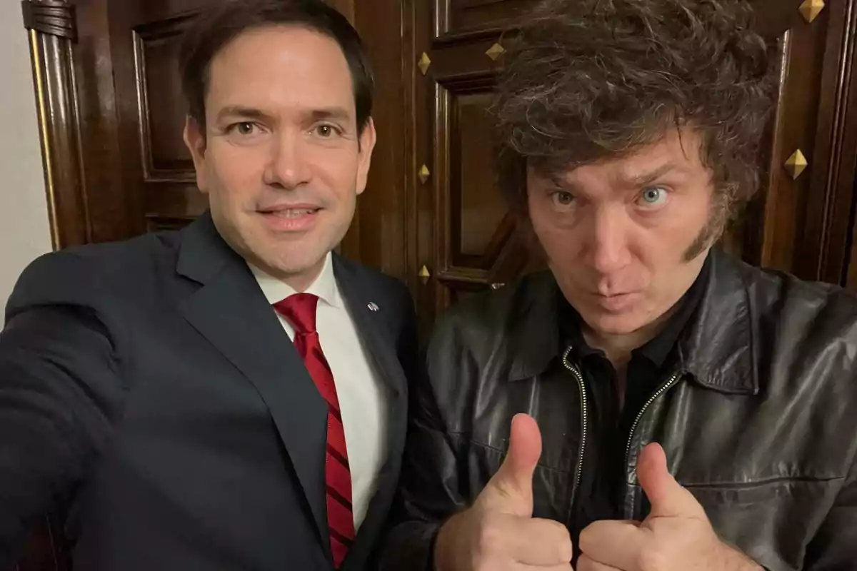 Dos personas posan para una foto, una de ellas lleva traje y corbata roja, mientras que la otra viste una chaqueta de cuero y muestra un gesto de aprobación con los pulgares hacia arriba. Dos personas posan para una foto, una de ellas lleva traje y corbata roja, mientras que la otra viste una chaqueta de cuero y muestra un gesto de aprobación con los pulgares hacia arriba.