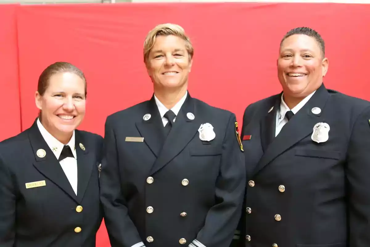 Tres personas en uniformes de bomberos posan sonriendo frente a un fondo rojo.