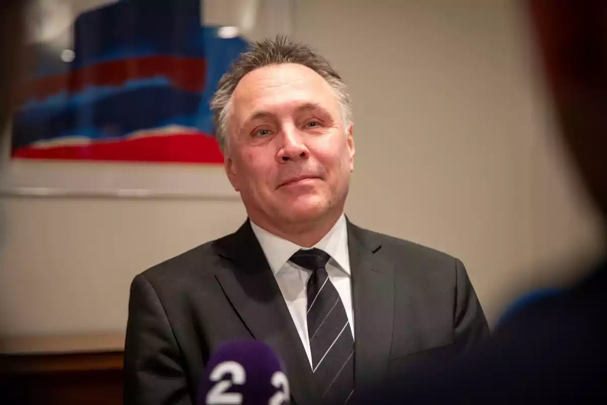 A man in a dark suit and tie is standing in front of a microphone in a room with an abstract painting on the wall. A man in a dark suit and tie is standing in front of a microphone in a room with an abstract painting on the wall.